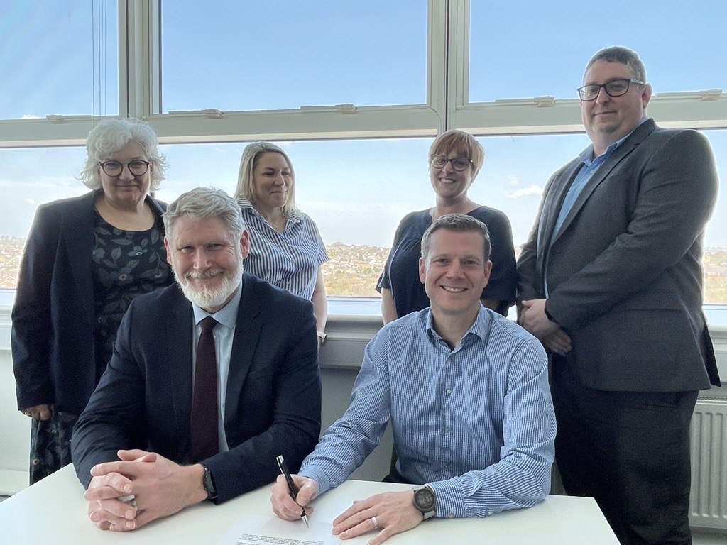 Two people sitting at desk signing a document with four people behind them, smiling